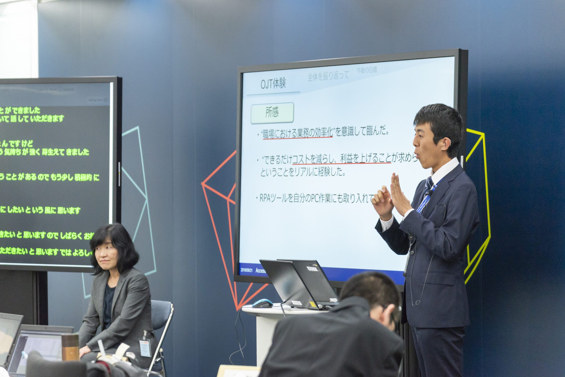 パワーポイントのスライドを写したディズプレイの横に立ち、手話を使いながらプレゼンテーションをする男性