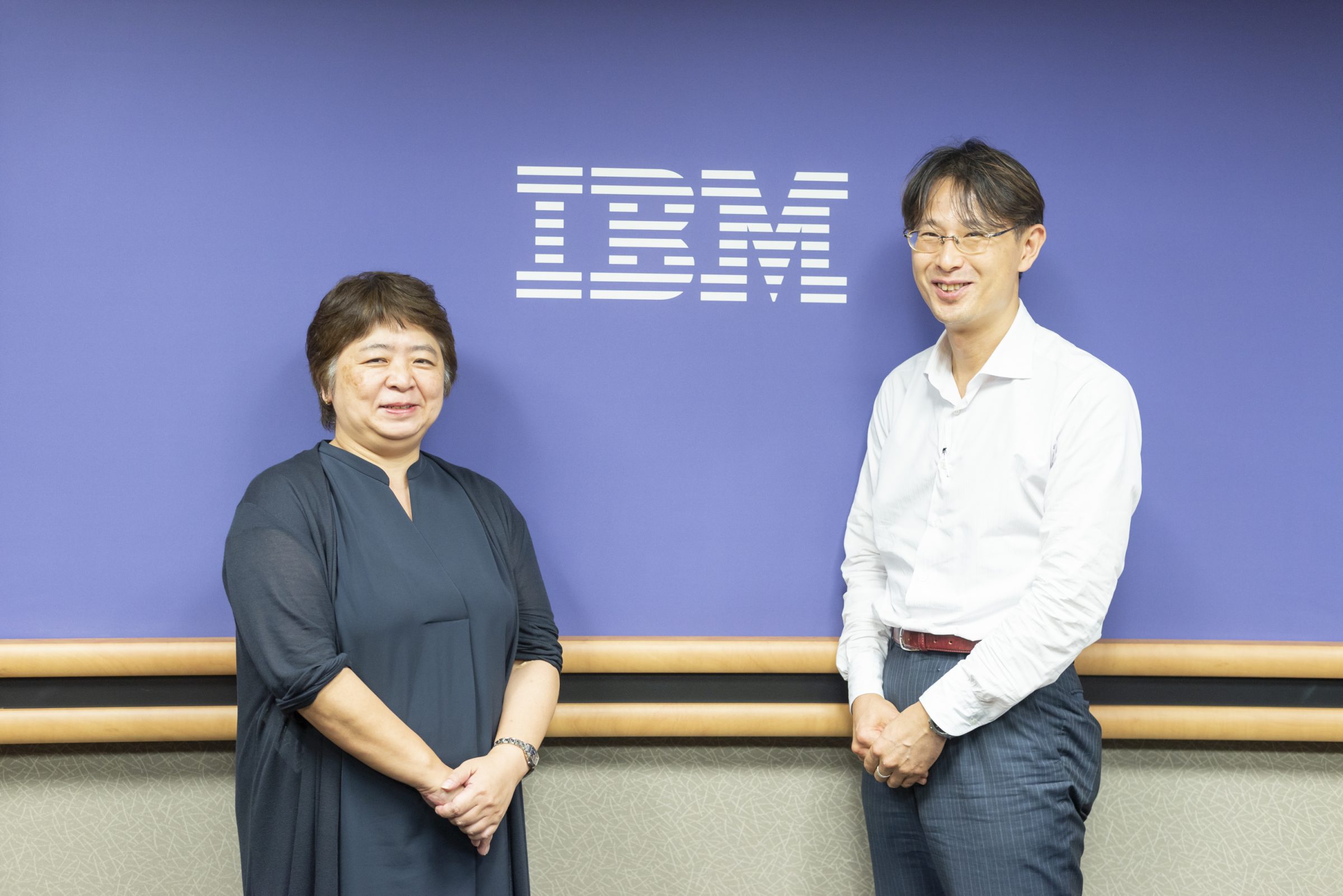 IBMのロゴが掲げられている青い壁の前で、ロゴを挟んで左に紺の服を着た女性、右に白いYシャツの男性が立っている