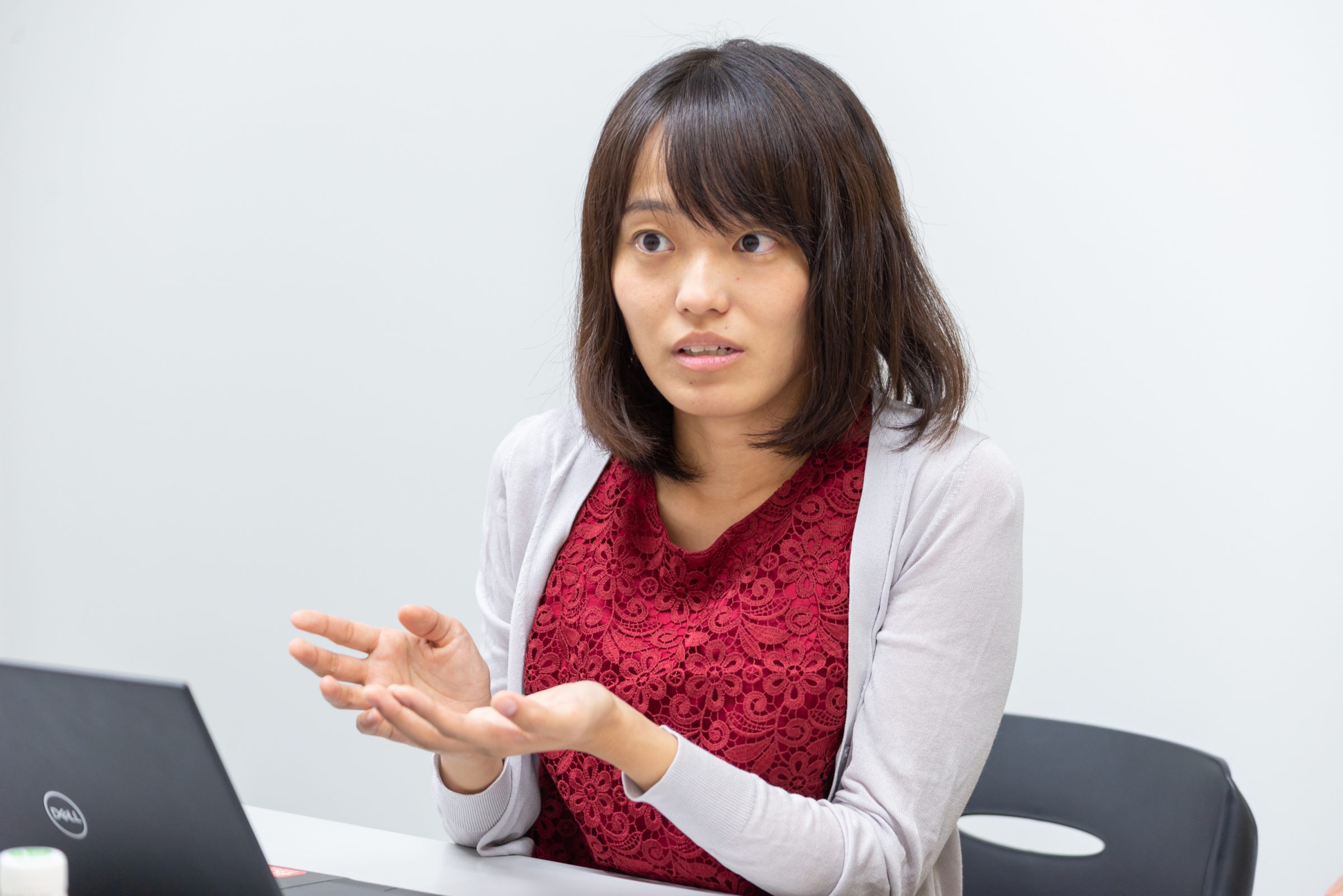 赤いレースの服にグレーのカーディガンを着たミディアムヘアの女性が手を前に出して語っている